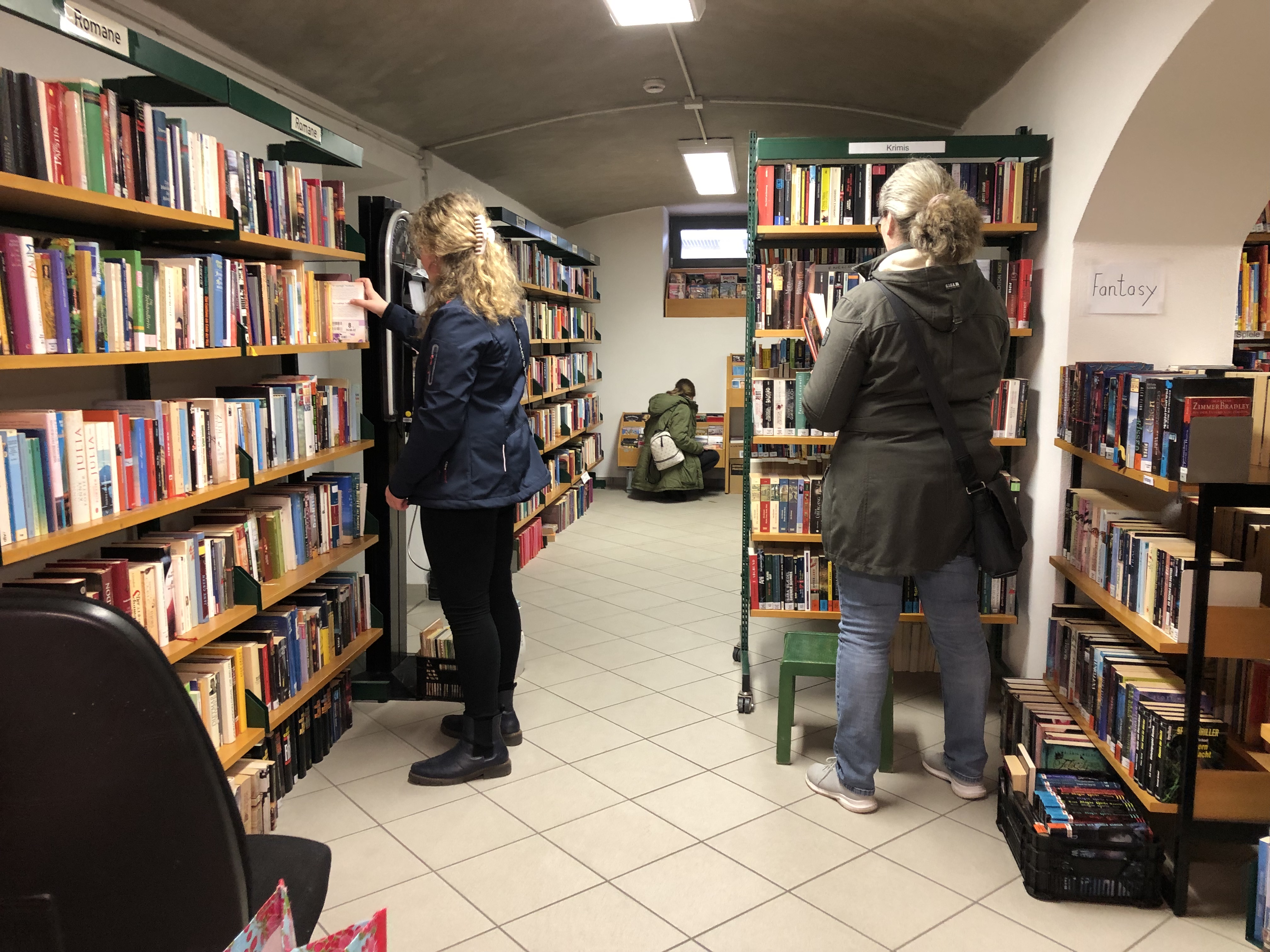 Im Gewölbekeller der Stadtbücherei stehen viele Bücherregale. Rechts und links eines Ganges sind zwei Frauen zu sehen, die mit dem Rücken zu Kamera ein Buch begutachten. Am Ende des Ganges kniet eine Frau vor einem Trog mit Büchern