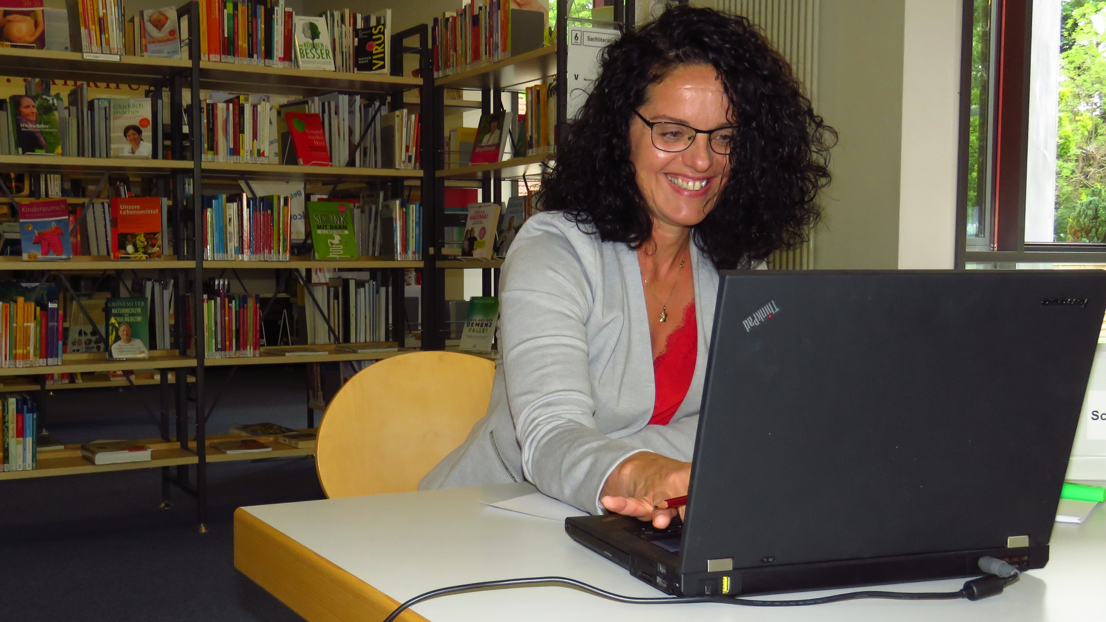 Eine dunkelhaarige Frau arbeitet lächelnd an ihrem Laptop an einem der Arbeitstische in der Stadtbücherei Wolfenbüttel