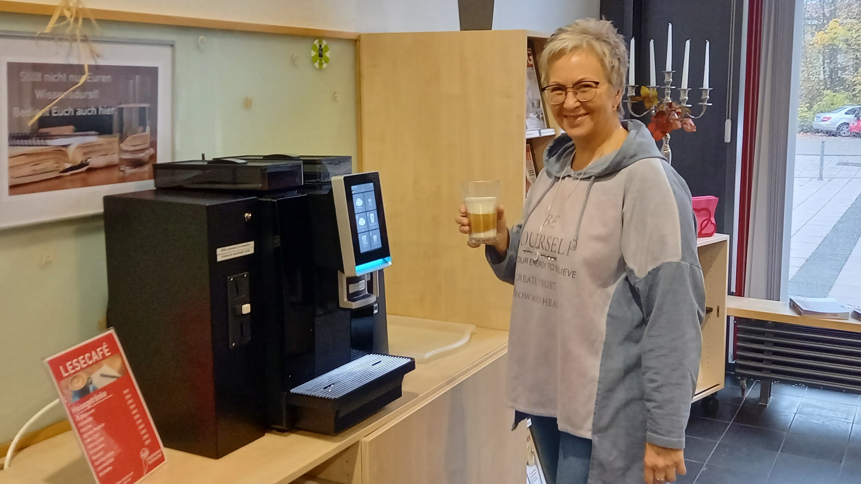 Eine Frau mit kurzen Haaren steht in der Stadtbücherei Wolfenbüttel vor dem Kaffeeautomaten. Sie hält ein Glas mit einem Latte Macchiato in der Hand und lächelt in die Kamera.